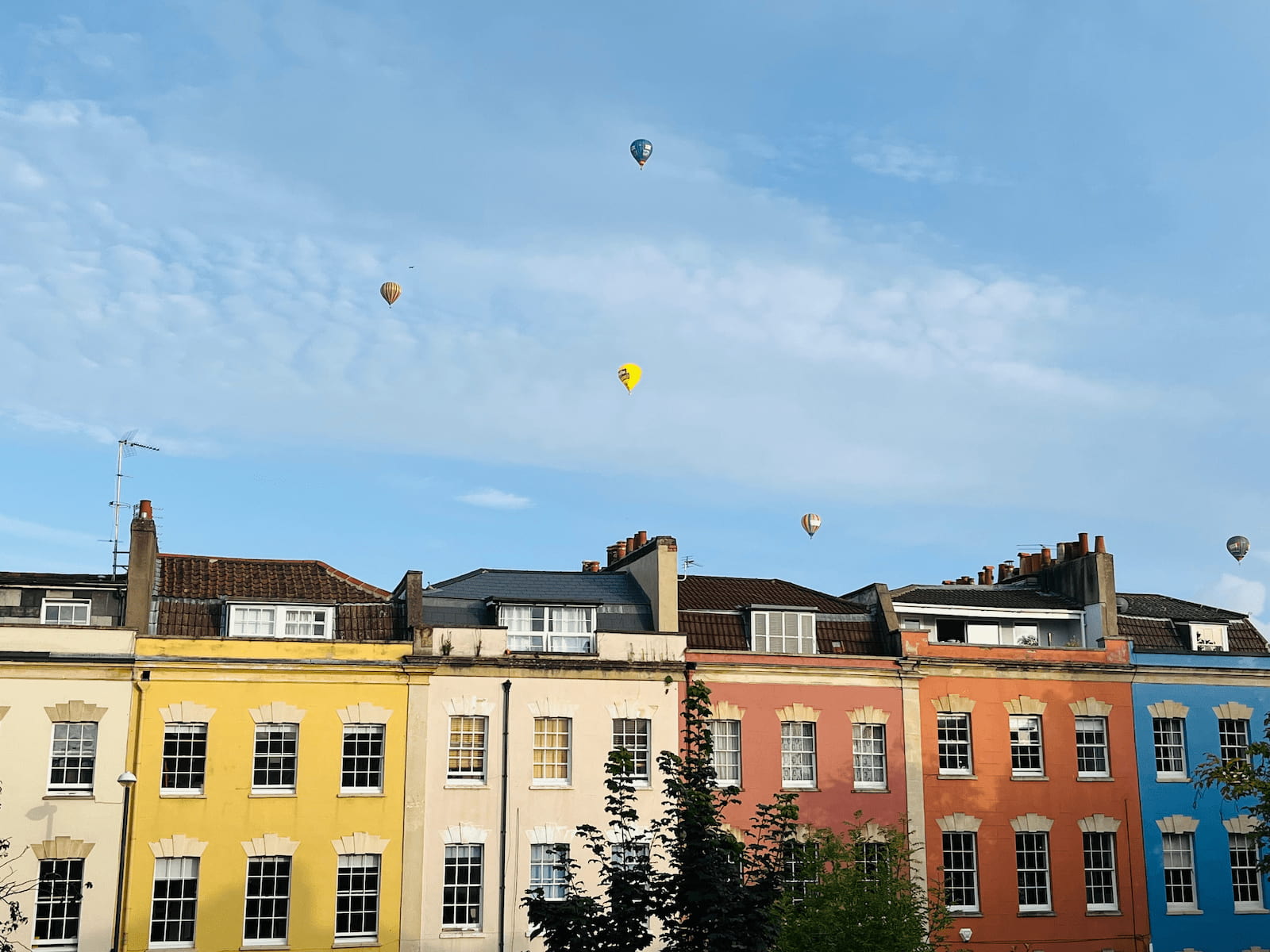 houses in various bright colors like yellow, blue and red, with a blue sky that contains 5 hot air balloons