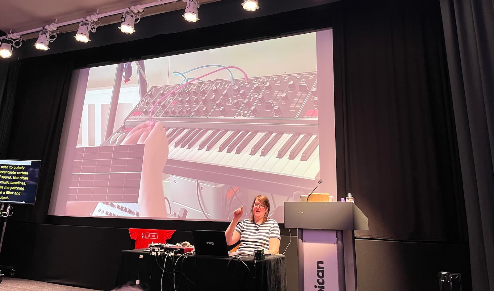 Katie behind a desk full of music tech, on the screen a video that shows cables on a synthesiser